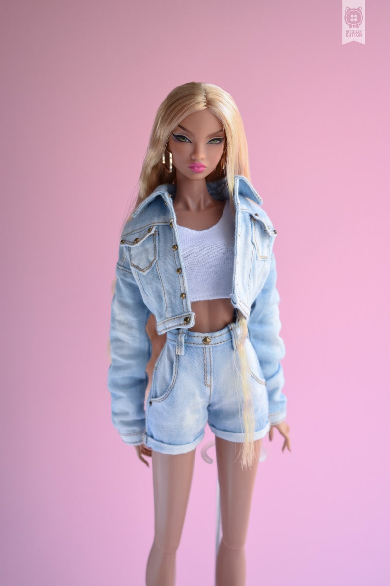May include: A fashion doll with long blonde hair wearing a light blue denim jacket and matching denim shorts. The doll has brown eyes and is wearing gold earrings.