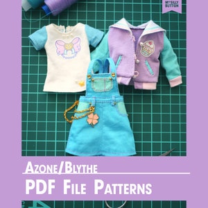 May include: A set of PDF sewing patterns for doll clothes. The patterns are for a doll wearing a white t-shirt with a pink bow, a blue denim overall dress, and a purple and blue jacket. The patterns are on a green cutting mat with a ruler and scissors.