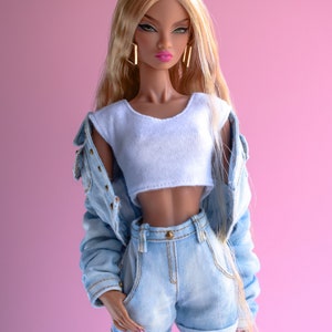 May include: A fashion doll with long blonde hair wearing a white crop top and light blue denim shorts with a denim jacket. The doll has brown skin and is wearing gold earrings.