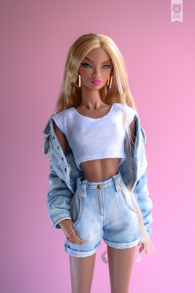 May include: A fashion doll with long blonde hair wearing a white crop top and light blue denim shorts with a denim jacket. The doll has brown skin and is wearing gold earrings.