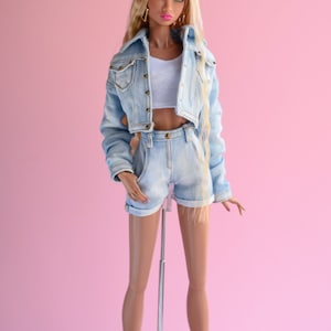 May include: A fashion doll wearing a light blue denim jacket and matching denim shorts. The doll has long blonde hair and is wearing a white tank top. The doll is standing on a silver stand against a pink background.