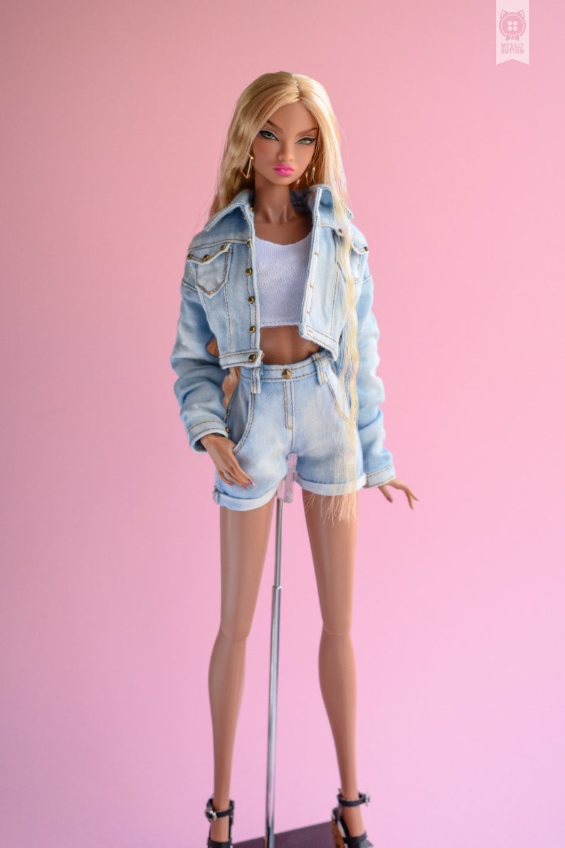May include: A fashion doll wearing a light blue denim jacket and matching denim shorts. The doll has long blonde hair and is wearing a white tank top. The doll is standing on a silver stand against a pink background.