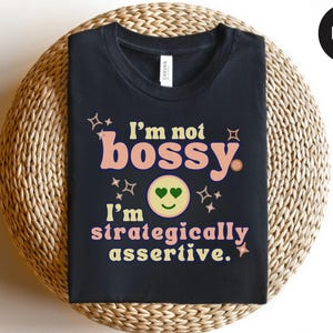 May include: Black t-shirt with the text "I'm not bossy, I'm strategically assertive." The design includes a smiley face with heart eyes and star accents. The shirt is displayed on a woven surface.