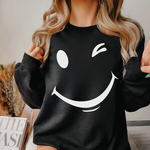 May include: Black sweatshirt with a white winking smiley face graphic.