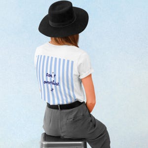 May include: A white t-shirt featuring a light blue and white vertical striped panel on the back with the dark blue text Don't overthink it, paired with a black wide-brimmed hat and dark grey trousers, viewed from the back against a light blue background.