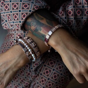 May include: A woman's wrist adorned with a stack of bracelets. The top bracelet is a rose gold chain with square-shaped charms in shades of pink and white. The other bracelets are a brown leather cord with a silver charm, a pearl strand, and a brown leather cord with a silver charm.