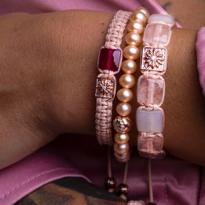 May include: A set of three bracelets with pink and white braided cords, pearl beads, and square-shaped charms. The charms feature a floral design and a red gemstone.