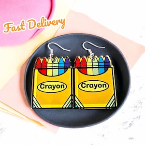 Peut inclure: Une paire de boucles d'oreilles en forme de boîte de crayons de couleur. Chaque boucle d'oreille représente une boîte de crayons avec un arc-en-ciel de couleurs et le mot "Crayon" imprimé sur le devant.