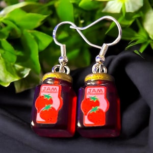 Op de afbeelding: Een paar zilveren oorhangers met een glazen pot charme. De potten zijn gevuld met een rode substantie en hebben een label met de tekst "JAM STRAWBERRY".