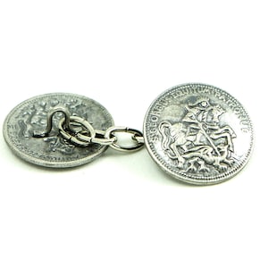 Könnte beinhalten: Silberfarbene Manschettenknöpfe mit einem detaillierten Design eines Ritters zu Pferd, verbunden durch eine kleine Kette. Die Manschettenknöpfe haben ein antikes Aussehen mit aufwendigen Details auf den kreisförmigen Flächen. Der Hintergrund ist schlicht weiß.