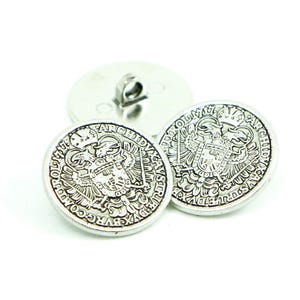 Può includere: Due bottoni argentati con un designo ornato che presenta un'aquila coronata e testo in una lingua straniera. I bottoni sono rotondi e hanno un bordo rialzato.