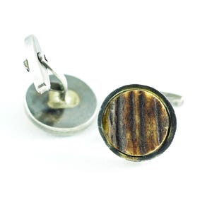 Pode incluir: Um par de abotoaduras circulares com um encaixe texturizado, castanho e estriado, inserido numa moldura de metal escuro. As abotoaduras possuem um mecanismo de fecho giratório prateado na parte traseira para fixar nas casas dos punhos.