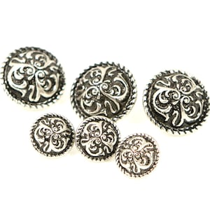 Könnte beinhalten: Fünf silberne Metallknöpfe mit einem dekorativen floralen Design. Die Knöpfe haben eine erhabene, strukturierte Oberfläche und eine abgerundete Form.