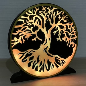 Peut inclure: Une décoration murale ronde en bois avec un fond noir et un motif d'arbre de vie doré. L'arbre est éclairé par derrière, créant une lueur chaude.