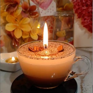 May include: A coffee-themed candle in a clear glass teacup. The candle has a layered appearance, with a dark brown base and a lighter top, sprinkled with cocoa powder and coffee beans. A lit wick provides a warm glow.