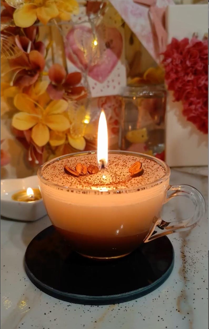 May include: A coffee-themed candle in a clear glass teacup. The candle has a layered appearance, with a dark brown base and a lighter top, sprinkled with cocoa powder and coffee beans. A lit wick provides a warm glow.