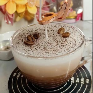 May include: A clear glass cup filled with layers of coffee and cream, topped with a white foam and sprinkled with ground coffee and whole coffee beans. A copper-colored candle holder is positioned above the cup, with a lit candle. The cup sits on a black coaster.