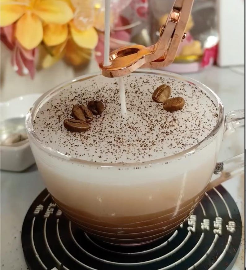 May include: A clear glass cup filled with layers of coffee and cream, topped with a white foam and sprinkled with ground coffee and whole coffee beans. A copper-colored candle holder is positioned above the cup, with a lit candle. The cup sits on a black coaster.