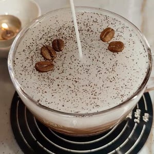 May include: A clear glass mug filled with a layered coffee drink, topped with a white foam and sprinkled with dark coffee grounds and whole coffee beans. A thin stream of liquid is being poured into the drink. A small candle and bowl are in the background.