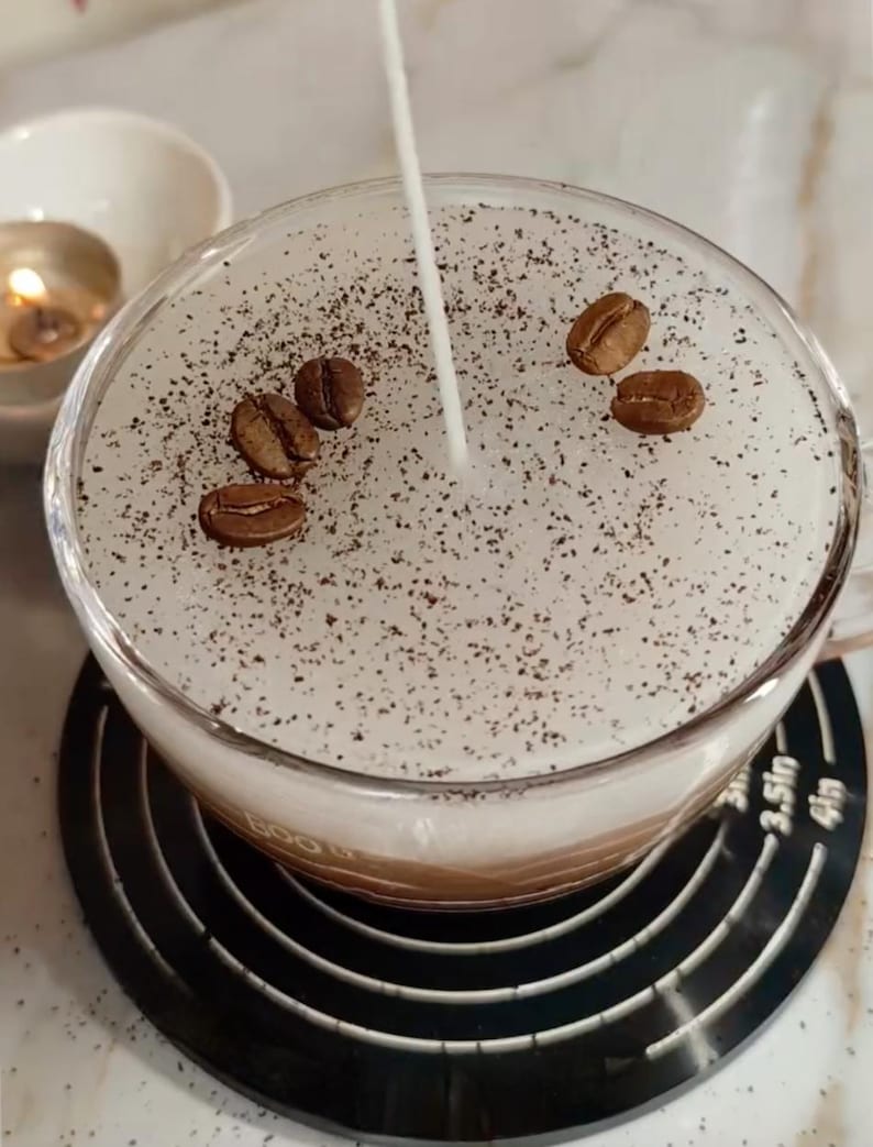 May include: A clear glass mug filled with a layered coffee drink, topped with a white foam and sprinkled with dark coffee grounds and whole coffee beans. A thin stream of liquid is being poured into the drink. A small candle and bowl are in the background.