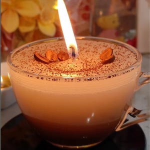 May include: A lit coffee-scented candle in a clear glass teacup. The candle has a layered appearance, with a dark brown base, a light brown middle, and a white top. Coffee beans are placed on top of the candle. The candle is on a black coaster.