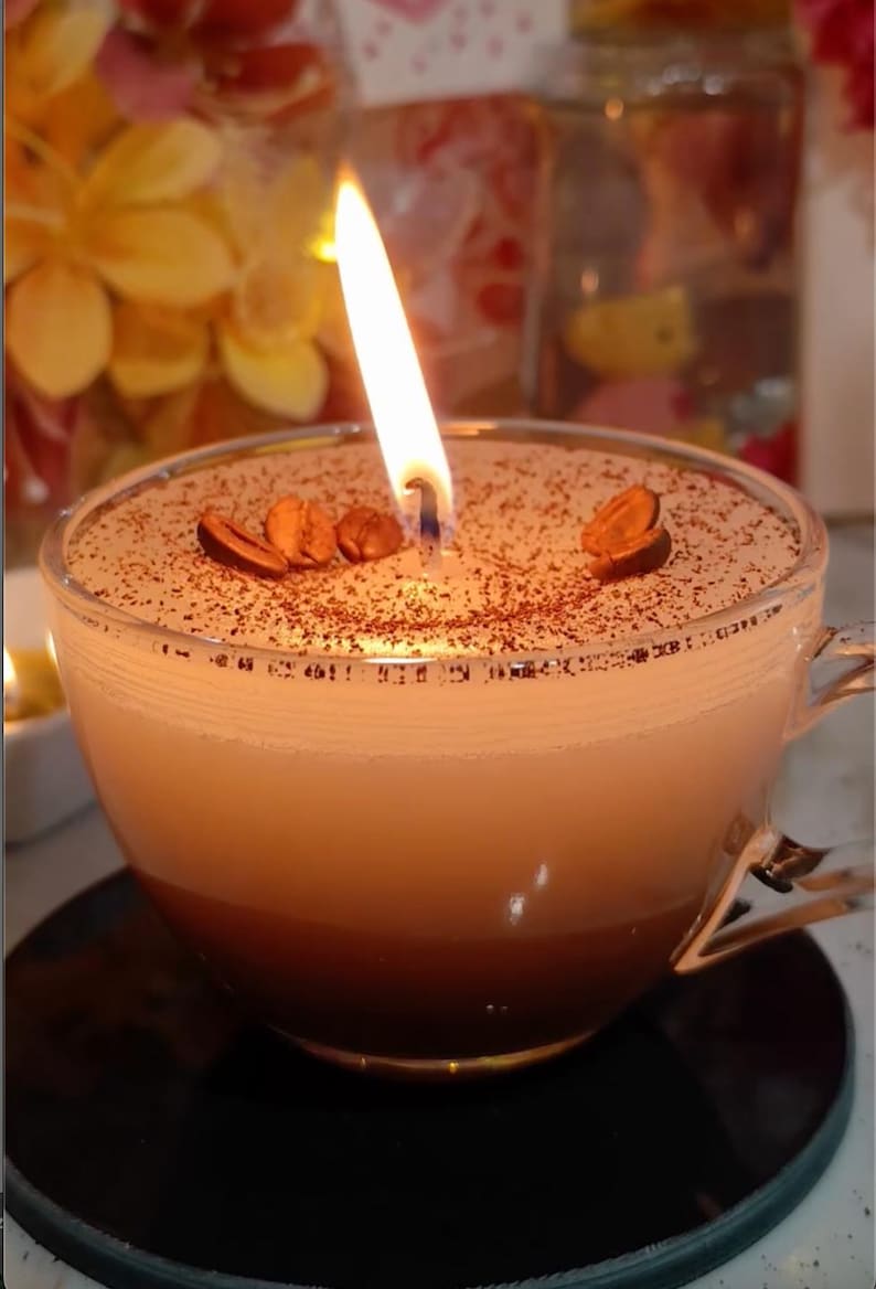 May include: A lit coffee-scented candle in a clear glass teacup. The candle has a layered appearance, with a dark brown base, a light brown middle, and a white top. Coffee beans are placed on top of the candle. The candle is on a black coaster.