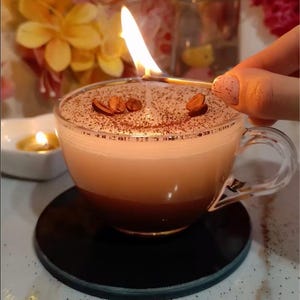 May include: A lit candle in a clear glass coffee cup. The candle has a layered appearance with a dark brown base, a light brown middle, and a white top. Coffee beans are placed on top. The cup sits on a black coaster.