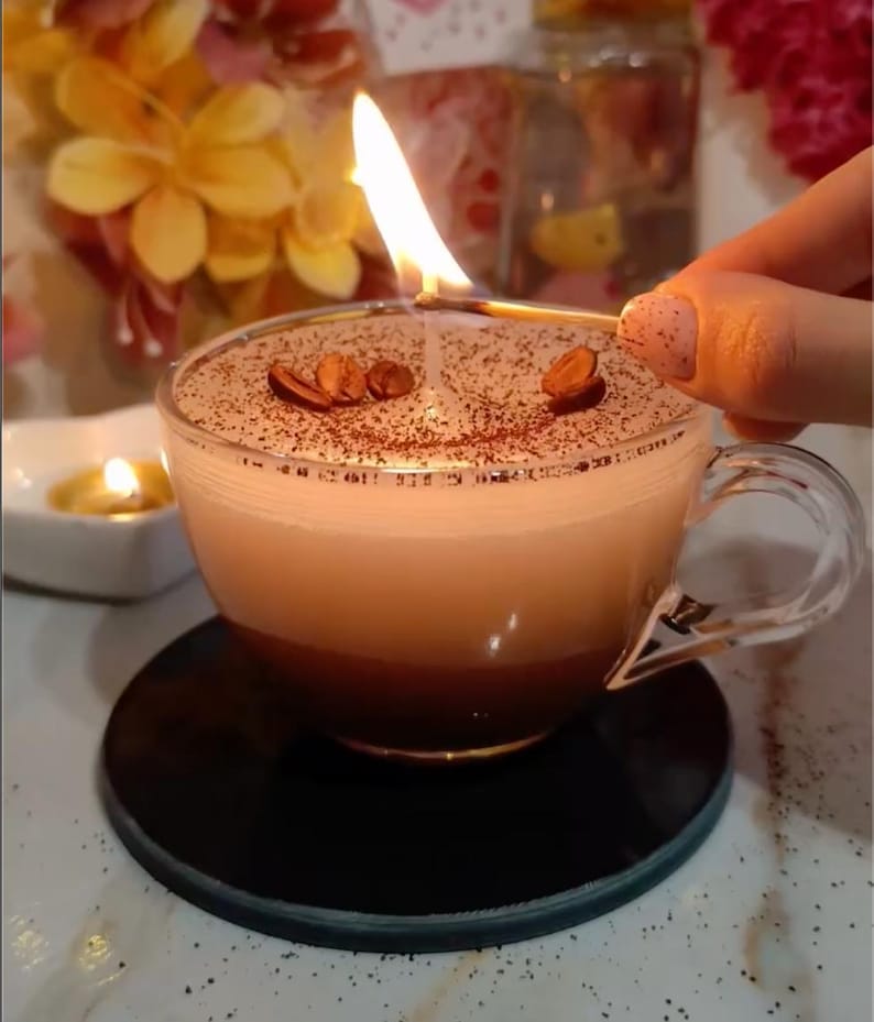 May include: A lit candle in a clear glass coffee cup. The candle has a layered appearance with a dark brown base, a light brown middle, and a white top. Coffee beans are placed on top. The cup sits on a black coaster.