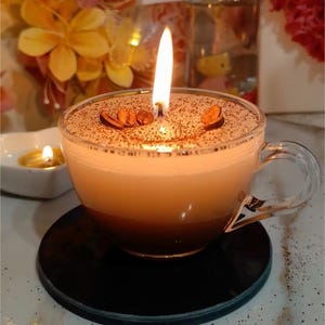 May include: A lit candle in a clear glass coffee cup, resembling a cappuccino. The candle has a brown, textured top with coffee bean decorations. The cup sits on a black coaster, with a blurred background of flowers and other candles.