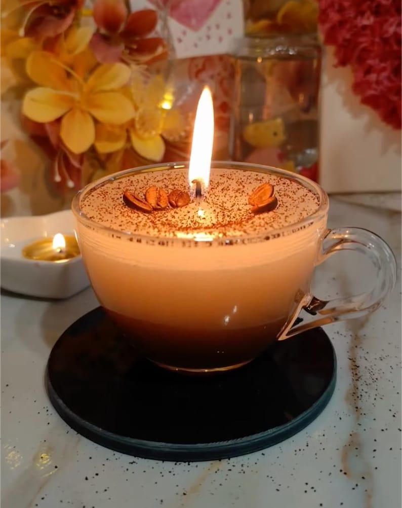 May include: A lit candle in a clear glass coffee cup, resembling a cappuccino. The candle has a brown, textured top with coffee bean decorations. The cup sits on a black coaster, with a blurred background of flowers and other candles.