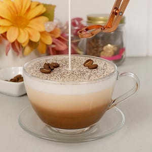 Peut inclure: Une tasse à café en verre transparent remplie d'une boisson en couches, garnie de grains de café et d'une fine poudre. De la crème est versée dans la tasse. Un petit bol blanc avec des grains de café et un bouquet de fleurs en arrière-plan.