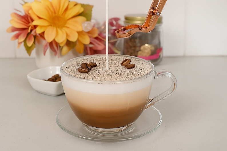 May include: A clear glass coffee cup with a saucer, filled with a layered beverage and topped with coffee beans. Cream is being poured into the cup. A small white bowl with coffee beans and a bouquet of flowers are in the background.