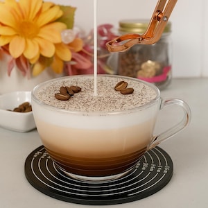 May include: A clear glass coffee mug filled with a layered latte, topped with coffee grounds and coffee beans. Cream is being poured into the coffee. The mug sits on a black coaster with measurement markings. A small white bowl with coffee beans is nearby.