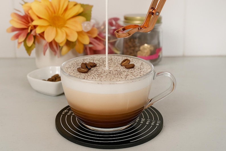 May include: A clear glass coffee mug filled with a layered latte, topped with coffee grounds and coffee beans. Cream is being poured into the coffee. The mug sits on a black coaster with measurement markings. A small white bowl with coffee beans is nearby.