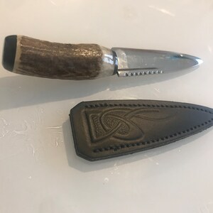 Peut inclure: Un petit couteau décoratif avec une lame en acier poli et un manche en corne. Le couteau a un bord dentelé et est accompagné d'un étui en cuir noir avec un motif de nœud celtique en relief. Mesure environ 20 cm.