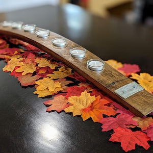 May include: A wooden candle holder with six glass votives, set on a bed of colorful autumn leaves. The holder is made from a curved piece of wood with metal accents, creating a rustic, seasonal centerpiece.