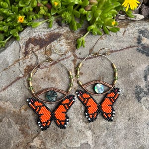 Puede incluir: Un par de pendientes de aro dorados con abalorios de mariposa naranja y un abalorio de ojo verde iridiscente. Los pendientes están sobre un fondo de roca marrón.