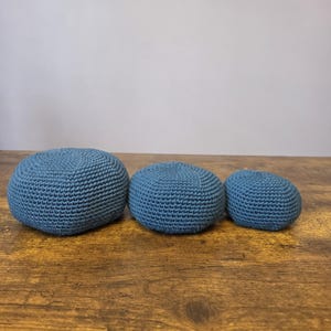 May include: Three blue crocheted beanbags of varying sizes are arranged on a wooden surface. The beanbags are a solid, muted blue color and have a textured, knitted appearance. The beanbags are round and appear to be filled with a soft material.