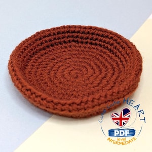 Pode incluir: Uma base para copos redonda de crochê, cor ferrugem, com um desenho em espiral. A base é feita à mão e tem uma borda ligeiramente elevada. A imagem também inclui um logotipo com um coração e o texto "PDF level Intermediate".