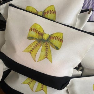 May include: Three white canvas zippered pouches with black trim. Each pouch features a yellow and red softball-themed bow.