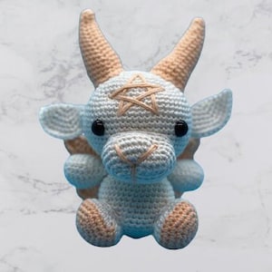 Peut inclure: Une chèvre en peluche crochetée, blanche et beige. La chèvre a des yeux noirs, des cornes beiges et un pentagramme beige cousu sur le front. Elle est assise, les pattes en avant, avec de petites oreilles arrondies. Jouet artisanal.