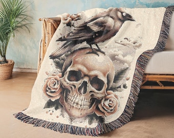 May include: A woven tapestry throw blanket features a skull with roses and a crow perched on top. The background is a cream color with a faded blue sky.