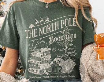 May include: A green t-shirt with a graphic print of a stack of books, a Christmas tree, and the text "The North Pole Book Club". The text below the graphic reads "Hot Cocoa, Sleigh Rides, Magical Reads".