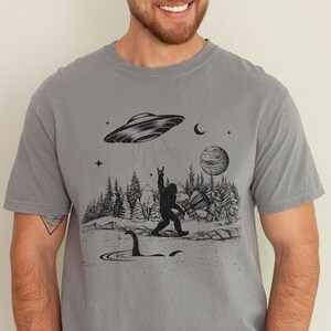 May include: A grey t-shirt featuring a black graphic print of a Bigfoot playing disc golf with a UFO flying overhead. The graphic also includes a Loch Ness Monster in the water.