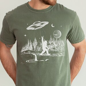 May include: A green t-shirt featuring a white graphic of a Bigfoot figure with a raised fist, standing in front of a lake with a Loch Ness Monster. A UFO hovers above Bigfoot, with stars and a crescent moon in the background.