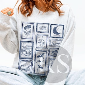 May include: A light gray sweatshirt featuring a collection of blue postage stamp designs. The stamps depict various illustrations, including flowers, books, a crescent moon with the word "Dreamer", and a dragon. The sweatshirt is a comfortable, casual style.