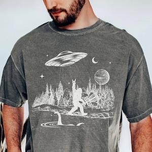 May include: A grey t-shirt with a white graphic print of a Bigfoot character holding a disc golf disc, a flying saucer, a moon, a planet, and a forest scene. The graphic also includes the text "Disc Golf"
