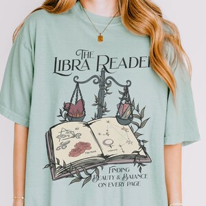 Pu&ograve; includere: Una maglietta verde chiaro con un design grafico che presenta un simbolo zodiacale della Bilancia, un libro e il testo "The Libra Reader: Finding Beauty & Balance on Every Page".