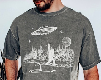 Könnte beinhalten: Ein graues T-Shirt mit einem weißen Grafikdruck eines Bigfoot-Charakters, der eine Discgolf-Scheibe hält, eine fliegende Untertasse, einen Mond, einen Planeten und eine Waldszene. Die Grafik enthält auch den Text "Disc Golf"