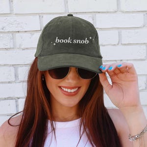 May include: A green corduroy baseball cap with white embroidery that reads "book snob" with small star accents.